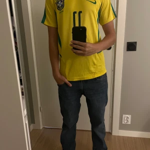 Brasilien Ronaldinho 11 Nike fotbollströja - Gul Brasilien fotbollströja med gröna detaljer från Nike, med Ronaldinho och nummer 11 på ryggen. Klassisk design med korta ärmar, rund hals och Brasilien-emblem på bröstet. Tillverkad i lätt och ventilerande polyester, perfekt för match eller träning.