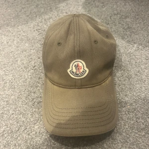 Olivgrön Moncler keps i bomull - Snygg olivgrön keps från Moncler med klassisk böjd skärm och broderad logotyp framtill. Justerbar rem baktill med diskret röd och vit detalj. Tillverkad i slitstark bomull, perfekt för dig som gillar stilrena accessoarer. Den är solblekt som man ser på bilden! Men utöver det är det inget slitage eller liknande 