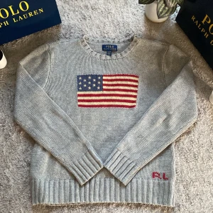 Grå stickad tröja Polo Ralph Lauren - Snygg grå stickad tröja från Polo Ralph Lauren med amerikansk flagga broderad på bröstet och RL-detalj nertill. Tröjan har rund halsringning, ribbade muddar och lång ärm. Perfekt för dig som gillar klassisk och stilren design.