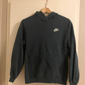 Grönblå Nike hoodie med logga - Säljer en grönblå hoodie från Nike med klassisk vit logga på bröstet. Tröjan har huva, magficka och långa ärmar. Perfekt för dig som gillar en enkel och stilren look. Materialet är mjukt och skönt, troligtvis bomullsmix. Storlek L (barn) / 147-158cm