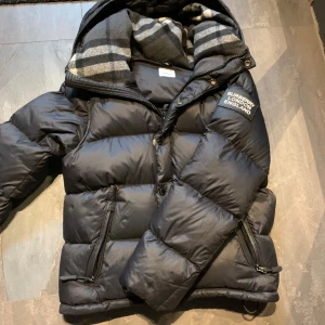 Burberry jacka  - Snygg svart pufferjacka från Burberry med tydlig logga på bröstet. Jackan har dragkedjor på fickorna, huva och ett klassiskt rutigt foder. Perfekt för kalla dagar och ger en clean och modern look.