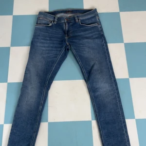 Blå jeans från Nudie Jeans, 33/32 - Säljer ett par klassiska blå jeans från Nudie Jeans med snygg tvätt och kontrastsömmar. Modellen har raka ben och fem fickor, samt Nudie-logga på bakfickan. Perfekta för dig som gillar stilrena och bekväma jeans.