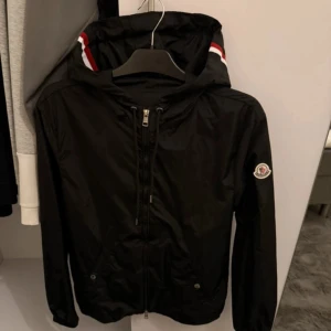 Moncler windbreaker - Svart vindjacka från Moncler med klassisk logga på ärmen och dragkedja framtill. Jackan har huva med röda och vita detaljer, samt praktiska fickor med knappar. Tillverkad i lätt polyester, perfekt för blåsiga dagar. Snygg och stilren design som lyfter din outfit.