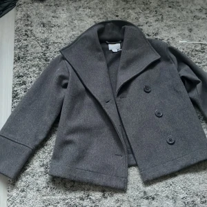 Grå kavajjacka från Gina Tricot - Snygg grå kavajjacka från Gina Tricot med bred krage och tre knappar framtill. Jackan är croppad och har raka, lite kortare ärmar. Perfekt för dig som vill ha en stilren och trendig look. Materialet känns mjukt och har en ullig känsla. Säljer pågrund av att jag växt ur den. Pris går att diskutera! 