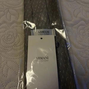 Armani Collezioni – ny slips i siden, originalförpackning - Elegant och exklusiv slips från Armani Collezioni, i klassisk design med diskret mönster. Perfekt accessoar för bröllop, fest eller affärsmöten.  Specifikationer:  Märke: Armani Collezioni  Färg: Grå med subtilt mönster  Material: 100% siden  Skick: Helt ny, aldrig använd  Förpackning: Fortfarande i originalförpackning med tagg kvar  Tillverkad i Italien