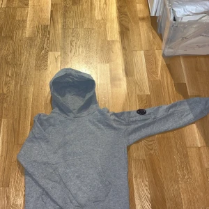 Grå hoodie från C.P. Company - Snygg grå hoodie från C.P. Company med klassisk huva och lång ärm. Ikonisk linsdetalj på ärmen och mjukt, fleeceliknande foder på insidan. Perfekt för dig som gillar streetwear och vill ha något bekvämt och stilrent. Köpt för 1500 använd ganska mycket