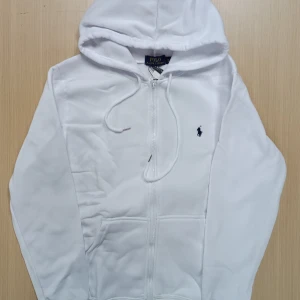 Vit hoodie från Polo Ralph Lauren - Vit hoodie från Polo Ralph Lauren med dragkedja framtill och klassisk huva med snörning. Tröjan har lång ärm, två fickor och den ikoniska broderade loggan på bröstet. Perfekt för en clean och stilren look.