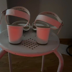 Vita sandaler med blockklack och spänne - Snygga vita sandaler från Clox med breda remmar och justerbart spänne med gulddetalj och liten hängande berlock. Skorna har blockklack och öppen tå, perfekt till sommarens outfits och fester. Tillverkade i syntetmaterial med mjuk innersula för extra komfort. Köpte för 500kr aldrig använda 