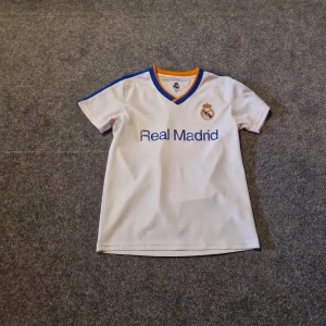 Real Madrid vit fotbollströja och shorts - Säljer ett Real Madrid fotbollskit med vit kortärmad tröja och matchande shorts. Tröjan har blå och orange detaljer vid krage och axlar, samt klubbmärke och Real Madrid-tryck på bröstet. Shortsen har blå sidopanel och klubbmärke. Materialet är lätt och andas, perfekt för träning.