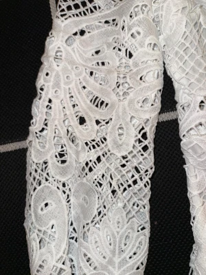 Vit spetsklännibg med volangdetaljer - Superfin vit klänning i spets med långärmad design och volangkant nedtill. Den har ett detaljerat broderat mönster och dragkedja i ryggen. Hög hals och genombrutna partier ger en lyxig och romantisk vibe. Perfekt för dig som gillar statement-plagg!