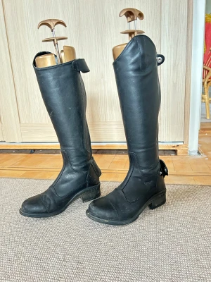 Svarta ridstövlar från CRW i skinn  - Säljer ett par svarta ridstövlar från CRW i äkta skinn. Stövlarna har rund tå, låg klack och dragkedja på sidan för enkel på- och avtagning. Klassisk design som passar perfekt till ridning och har en stabil platt sula. Änvänd lite. Nypris: 1200kr