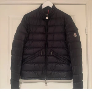 Svart Moncler pufferjacka herr - Svart pufferjacka från Moncler med klassisk logga på ärmen, ståkrage och två snedställda fickor med dragkedja. Jackan har quiltad design och är tillverkad i polyester med dunfyllning. Perfekt för kalla dagar och har en snygg, stilren look.