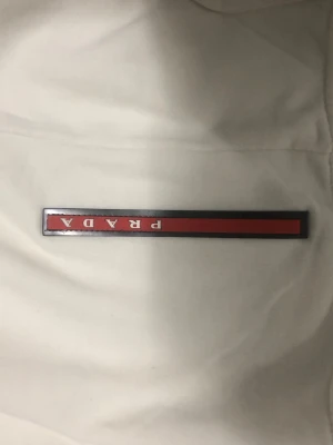 Vit hoodie från Prada med logga - Säljer en clean vit hoodie från Prada i storlek L. Tröjan har huva med snörning och den klassiska Prada-loggan broderad i rött och vitt på bröstet. Mjukt material och ribbade muddar vid ärmslut och nederkant. Perfekt för dig som gillar exklusiva streetwear-vibes.