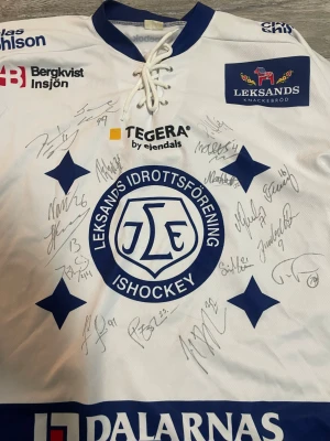 Leksands IF signerad hockeytröja - Säljer en vit och blå långärmad hockeytröja från Leksands IF, tillverkad av Reebok. Tröjan har klubbens logga på bröstet, flera sponsormärken och är signerad av spelare. Materialet är polyester och har snörning vid halsen samt blå detaljer och stjärnor.