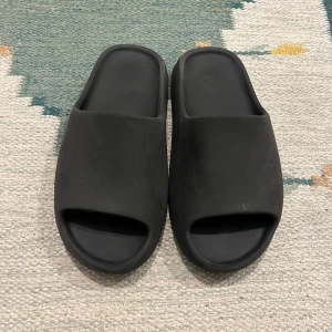 Svarta Adidas Yeezy slides - Svarta Adidas Yeezy slides med chunky, räfflad sula och öppen tå. Tillverkade i syntetmaterial för en mjuk och bekväm känsla. Minimalistisk design med Adidas-logga på fotbädden. Perfekta för chill dagar och enkel att slänga på sig.