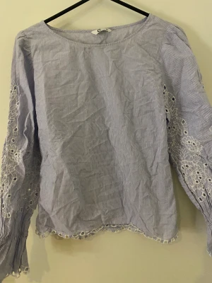 Blårandig blus med spetsdetaljer - Söt blårandig blus med långa ärmar och broderade spetsdetaljer längs ärmar och nederkant. Tunt bomullstyg och rund halsringning. Perfekt för dig som gillar romantiska och feminina detaljer.