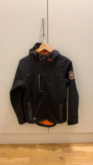 Helly Hansen vindjacka  - Svart vindjacka från Helly Hansen Workwear med orange fleecefoder och coola detaljer i orange. Jackan har huva, dragkedja framtill och två fickor vid magen och en ficka vid bröstet. Storlek Xs  nypris 800kr.  Inga defekter. Skriv till mig om du har minsta fundering mvh Alex 