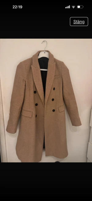 Beige kappa med svarta knappar - Snygg beige kappa med dubbelknäppning och svarta knappar. Jackan har klassisk krage, två fickor med lock och är helfodrad. Perfekt för dig som gillar stilrena och tidlösa plagg med lite längre passform.