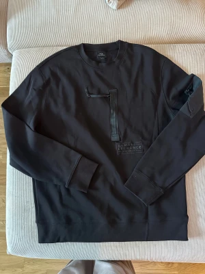 Svart sweatshirt från Armani Exchange - Svart sweatshirt från Armani Exchange med coolt band-detalj på bröstet och tryckt logga framtill. Tröjan har rund hals, långa ärmar och ribbade muddar. Materialet är mjuk bomull med inslag av polyester för extra komfort. Perfekt för dig som gillar stilrena och moderna plagg. Helt ny, julklapp förra året dock för stor M motsvarar mer L. Tröjan är enbart provad. Nypris 1890kr