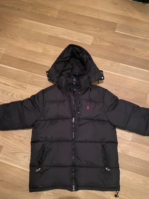 Svart dunjacka från Polo Ralph Lauren - Svart dunjacka från Polo Ralph Lauren med huva och röd logga på bröstet. Jackan har två sidofickor med dragkedja och en rejäl dragkedja framtill. Perfekt för kalla dagar och har en klassisk, sportig look.