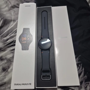 Galaxy Watch FE - Samsung Galaxy Watch FE i nyskick, komplett med originalförpackning och laddkabel. Modern design med svart armband och färgglada detaljer. Perfekt för träning, hälsa och smarta funktioner. Inga synliga repor eller skador, ser oanvänd ut.