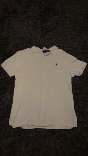 Vit pikétröja från Polo Ralph Lauren - Vit pikétröja från Polo Ralph Lauren med klassisk krage och två knappar framtill. Tröjan har korta ärmar och den ikoniska broderade loggan i marinblått på bröstet. Tillverkad i mjuk bomull som är skön mot huden.