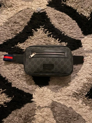 Gucci svart axelväska med monogram - Snygg svart axelväska från Gucci med klassiskt GG-monogram över hela väskan. Väskan har en silverfärgad dragkedja framtill och justerbart band i blått och rött. Materialet är en mix av skinn och syntet, perfekt för att bära det viktigaste.