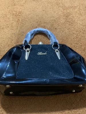 Svart glittrig handväska med gulddetaljer - Snygg svart handväska från Simuch med glittrig framsida och blanka sidor i syntetmaterial. Väskan har guldfärgade detaljer, dekorativa spännen och ett elegant mönstrat guldlås med stenar. Perfekt för dig som gillar glam och stil.