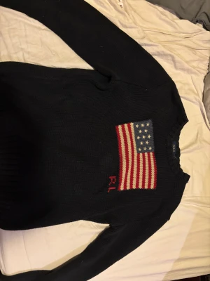 Svart stickad tröja Polo Ralph Lauren - Svart stickad tröja från Polo Ralph Lauren med amerikansk flagga i rött, vitt och blått på bröstet samt RL-broderi. Tröjan har rund hals och långa ärmar med ribbade muddar. Perfekt för dig som gillar klassisk och cool stil. Pris kan diskuteras.