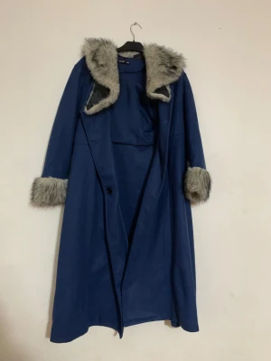 Blå kappa med fuskpälsdetaljer - Snygg mörkblå kappa från Vero Moda med fluffiga grå fuskpälsdetaljer på krage och ärmslut. Lång och loose fit, perfekt för kalla dagar. Stängs med knapp framtill och har en stilren look som sticker ut.