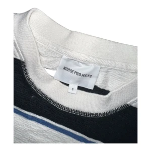 Randig t-shirt från Norse Projects - Snygg t-shirt från Norse Projects i storlek S med breda svarta och vita ränder samt tunna blå linjer. Klassisk rund hals och korta ärmar. Tillverkad i mjuk bomull som känns skön mot huden. Perfekt för dig som gillar stilrena och grafiska mönster.