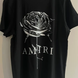 Svart Amiri t-shirt med tryck - Två svarta t-shirts från Amiri med coola tryck framtill. Ena har en stor silverfärgad ros och logga, den andra har Amiri-loggan med färgstänk som rinner ner. Båda har rund hals och korta ärmar, tillverkade i mjuk bomull. Perfekt för dig som gillar streetwear och snygga detaljer.