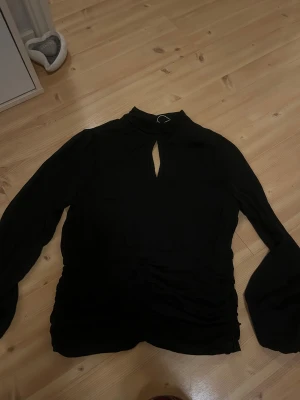 Svart långärmad blus - Svart blus med lång ärm och snygg nyckelhålsringning vid halsen. Toppen har rynkade detaljer längs midjan och en hög krage som ger en cool vibe. Materialet känns lätt och flowy, perfekt för dig som gillar stilrena plagg med twist.