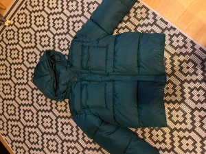 Mörkgrön dunjacka med huva - Säljer en mörkgrön pufferjacka med huva och stora fickor framtill. Jackan har quiltad design och en reflekterande detalj på ryggen. Perfekt för kalla dagar och har en loose passform som ger skön streetkänsla.