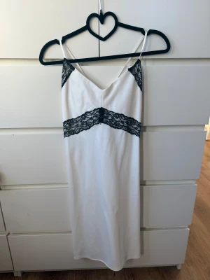 Vit klänning med svart spets från Kappahl - Supersnygg vit klänning från Kappahl med tunna axelband och svart spetsdetalj över byst och midja. Klänningen har snörning i ryggen och är i mjukt, glansigt material som ger en lyxig känsla. Perfekt för sommarkvällar när du vill känna dig extra fin.