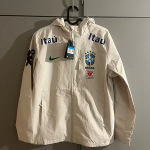 Beige Brasil Nike vindjacka M - Snygg beige vindjacka från Nike med Brasil-logga och flera tryck på bröstet och ärmarna. Jackan har huva, dragkedja framtill och meshfoder på insidan. Perfekt för dig som gillar fotboll och vill ha en sportig stil.
