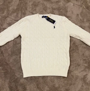 Vit kabelstickad tröja Polo Ralph Lauren - Snygg vit kabelstickad tröja från Polo Ralph Lauren med klassisk rund hals och svart broderad logga på bröstet. Tröjan har långärmad passform och ribbade muddar vid ärmslut och nederkant. Perfekt för dig som gillar stilrena och tidlösa plagg.