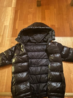 Svart pufferjacka från Alessandro - Svart pufferjacka från Alessandro med glansig finish och huva. Jackan har diskret logotyp på bröstet och ett emblem på ärmen. Fylld med syntetmaterial och har dragkedja framtill. Perfekt för kalla dagar och har en modern, oversized look.