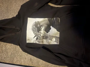 Svart Tupac hoodie med tryck - Svart hoodie med stort Tupac-tryck på bröstet. Tröjan har huva med snörning och en klassisk magficka. Perfekt för dig som gillar streetwear och vill ha en cool look med ikonisk hiphop-stil.