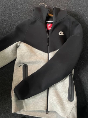 Nike hoodie i svart och grå - Nike hoodie med huva i svart och ljusgrått, med dragkedja framtill och två sidofickor med dragkedja. Snyggt broderad Nike-logga på bröstet. Perfekt för chill eller träning, och har en modern, sportig look.