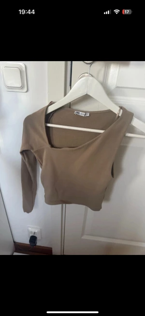 Beige oneshoulder topp från Zara - Säljer en beige oneshoulder topp från Zara med lång ärm på ena sidan och ärmlös på andra. Croppad passform och mjukt stretchigt material som sitter snyggt. Perfekt för dig som gillar trendiga och unika plagg med cool asymmetri.