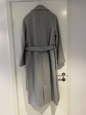 Grå lång kappa med bälte - Snygg grå design kappa i ullmix vilket gör den varm. Lång passform, två fickor på sidorna och slits bak. Den är från 2NDDAY och ordinarie pris är ca 3000! Den är använd max 3 gånger och har inga som helst tecken på användning! Bara att höra av sig för vilka frågor som helst!