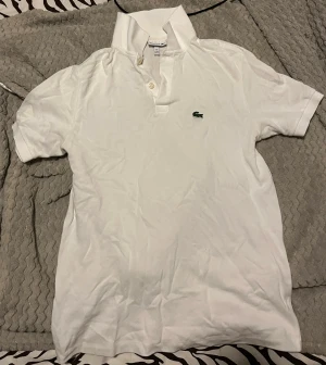 Vit Lacoste pikétröja med knappar - Vit pikétröja från Lacoste med klassisk krage och två knappar framtill. Tröjan har det ikoniska gröna krokodilmärket broderat på bröstet. Tillverkad i mjuk bomull som är skön mot huden och har korta ärmar. Perfekt för en clean och sportig look. Använd en gång tidigare utöver det är den orörd.
