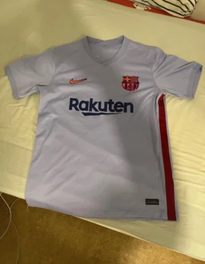 Barcelona fotbollströja Nike ljusblå - Barcelona fotbollströja från Nike i ljusblå färg med röd detalj längs sidan. Tröjan har korta ärmar, V-ringad hals och tryckt Rakuten-logga på bröstet. FCB-emblemet och Nike-loggan är i metallic orange. Materialet är lätt och andas, perfekt för match eller träning.