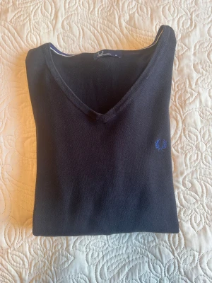 Svart v-ringad tröja Fred Perry XL - Svart v-ringad tröja från Fred Perry i storlek XL. Tröjan har lång ärm, ribbade muddar och blå broderad logga på bröstet. Materialet är mjukt och tunt, perfekt för lager-på-lager.  Jag upplever den svart men man kan nog också se den som mörk marinblå. 