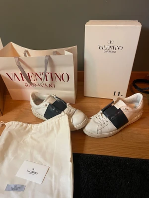 Valentino Garavani Open sneakers vit/blå - Säljer ett par snygga Valentino Garavani Open sneakers i vitt skinn med bred blått läderdetalj över vristen. Klassisk rund tå, platt sula och snörning framtill. Kommer med originalkartong, dustbag och påse. Perfekta för dig som gillar lyxiga och stilrena sneakers.