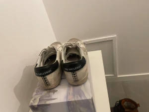 Vita sneakers från The Gallerist - Säljer ett par vita sneakers från The Gallerist med svart hälparti och grå detalj på sidan i form av en klöver. Skorna har snörning, rund tå och platt sula. Ovandelen är i skinn och de har en klassisk, stilren look som passar till det mesta.