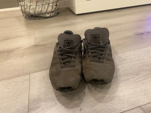 Adidas LA Trainer grå sneakers - Säljer ett par Adidas LA Trainer sneakers i mörkgrå färg med klassisk snörning och rund tå. Skorna har en textil ovandel med diskret struktur och tre dämpande pluggar i sulan. Ikonisk Adidas-logga på plösen och hälen. Perfekt för dig som gillar stilrena och sportiga sneakers.