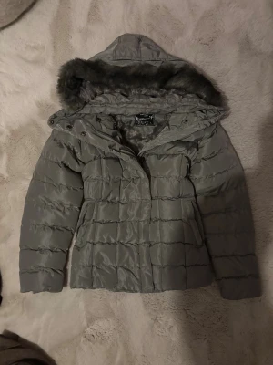 Grå khaki pufferjacka med fuskpäls - Grå pufferjacka från Nature med fluffigt fuskpälsfoder och huva med pälsdetalj. Jackan har quiltad design, stora knappar framtill och dragkedja vid fickorna. Perfekt för kalla dagar och har en snygg, lite längre passform.