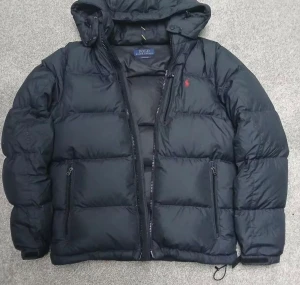 Svart dunjacka Polo Ralph Lauren M - Svart dunjacka från Polo Ralph Lauren med huva och röd logga på bröstet. Jackan har dragkedja framtill, två sidofickor med dragkedja och quiltad design. Materialet är bomull och syntet, perfekt för kalla dagar. Snygg och klassisk look med normal passform.